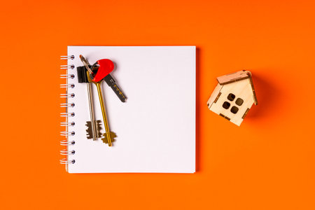 A empty notebook on a spiral, house keys and a wooden house on an orange background. Top view, real estate purchase conceptの写真素材