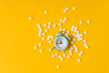 The alarm clock is lying on a yellow background in the middle of the scattered pills. The concept of...