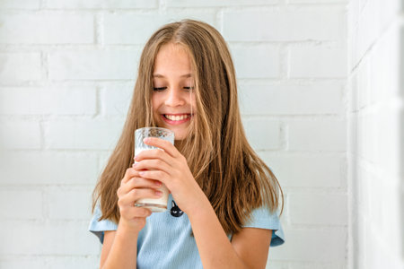 A beautiful little girl is having fun drinking a glass of fresh milk against a white brick wallの写真素材