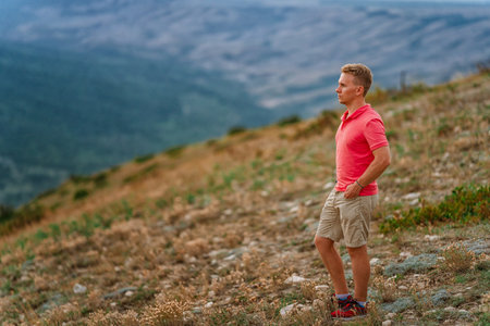 A young blond man in a pink shirt conquered the top of the mountain at sunset. Beautiful mountain landscape with a gradient. The concept of enjoying nature.の写真素材