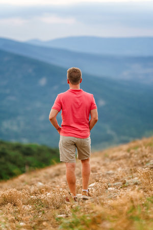 A young blond man in a pink shirt conquered the top of the mountain at sunset. Beautiful mountain landscape with a gradient. The concept of enjoying nature.の写真素材