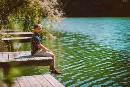 A young man is resting on a wooden pier, relaxing enjoying the scenery of a forest and a mountain lake.の写真素材