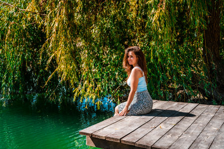 A young beautiful woman is sitting on a wooden pier on a delightful azure mountain lake surrounded by forestの写真素材