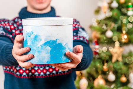 A young man holds a Christmas gift to the camera against the background of a decorated Christmas tree. Celebrating Merry Christmas or new Year at home.の写真素材