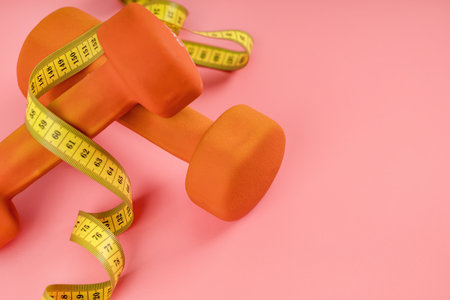 Dumbbells with a yellow measuring tape on a pink background. The concept of sports, diet and weight lossの写真素材