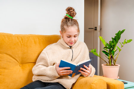 A little teenage girl with glasses is sitting on a yellow sofa at home and reading a book with a...