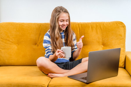 Smiling European teenage girl is using a laptop for video calls to friends and parents sitting on a yellow sofa. Distance learning online education.の写真素材