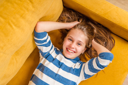 A close-up portrait of a smiling beautiful little blonde girl lying on a yellow sofa, a photo on top of herの写真素材