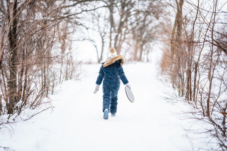 A little girl in a warm suit holds a tray for skating down an ice slide in winter. Winter fun in the snow.の写真素材