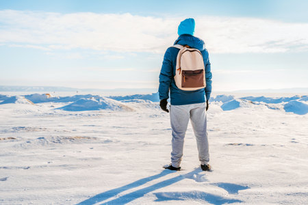 A lone man in a warm jacket and backpack stands in the middle of a snowy desert on a frozen river on a sunny winter dayの写真素材