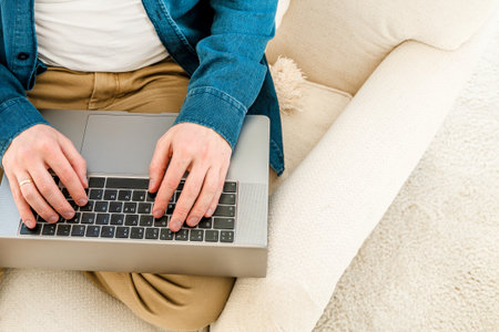 A young man in a blue shirt is working with a laptop while sitting on the couch at home in a cozy room. The concept of using the technology of the contact communication device business, IT technologの写真素材