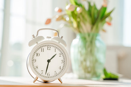 The alarm clock stands in a white modern sunny room on a wooden table, a bouquet of flowers in the backgroundの写真素材