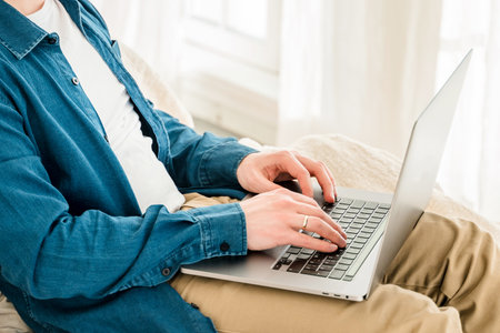 A young man in a blue shirt is working with a laptop while sitting on the couch at home in a cozy room. The concept of using the technology of the contact communication device business, IT technologの写真素材