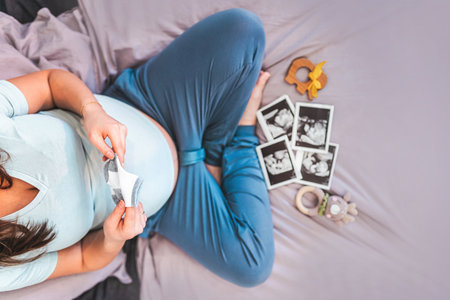 A pregnant woman is sitting on the bed and looking at ultrasound pictures of the baby, the concept of motherhood and preparation for the birth of a childの写真素材
