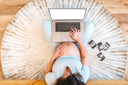 A pregnant woman is sitting on the carpet and using a laptop for shopping or work, ultrasound images of her baby are spread out on the carpetの写真素材