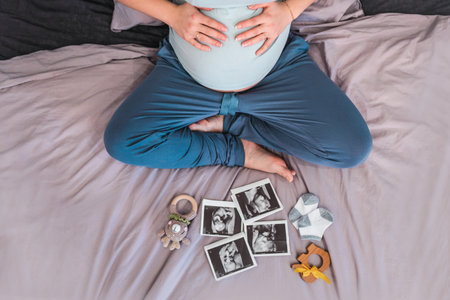 A pregnant woman is sitting on the bed and looking at ultrasound pictures of the baby, the concept of motherhood and preparation for the birth of a childの写真素材