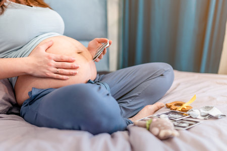 A pregnant woman is sitting on the bed and looking at ultrasound pictures of the baby, the concept of motherhood and preparation for the birth of a childの写真素材