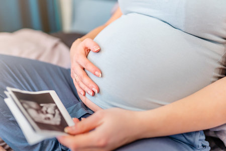 A pregnant woman is sitting on the bed and looking at ultrasound pictures of the baby, the concept of motherhood and preparation for the birth of a childの写真素材