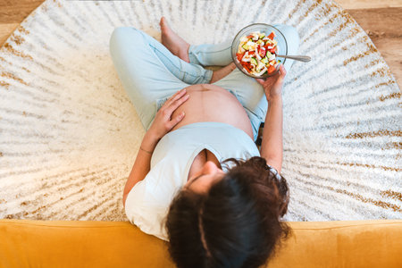 A pregnant woman holds a bowl of vegetable salad sitting on the carpet, top view, the concept of proper nutrition and diet in pregnant womenの写真素材