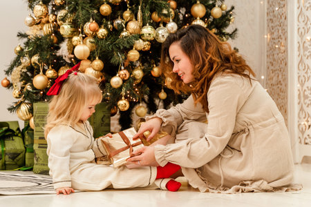 A young mother in a dress hands a gift to her 3-year-old daughter at a decorated Christmas tree. Home decorations for the New Year. Family Christmas traditions.の写真素材