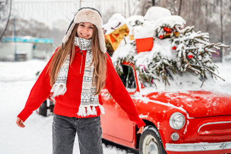 Beautiful girl in a red sweater, hat and scarf on the background of a red retro car with a Christmas tree.の写真素材