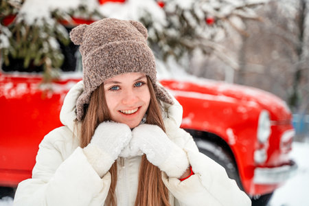 A beautiful young woman in a winter hat with earflaps next to a retro red car decorated for Christmas. A snowy winter day, christmas tree on car roof.の写真素材