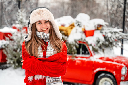 A beautiful young woman in a winter hat with earflaps next to a retro red car decorated for Christmas. A snowy winter day, christmas tree on car roof.の写真素材