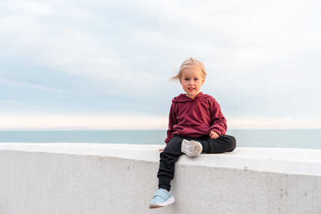 A cute child girl sits on a stone embankment against the background of a smooth line of sky and sea,...