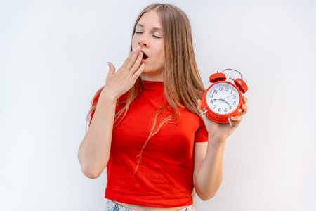 A beautiful young girl holds a red alarm clock and yawns against an isolated background.