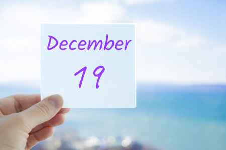 December 19th. Hand holding sticker with text December 19 on the blurred background of the sea and sky.の写真素材