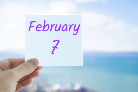 February 7th. Hand holding sticker with text February 7 on the blurred background of the sea and sky. Copy space for text. Month in calendar concept.の写真素材
