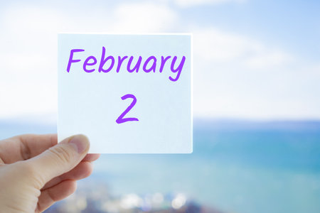 February 2nd. Hand holding sticker with text February 2 on the blurred background of the sea and sky. Copy space for text. Month in calendar concept.の写真素材