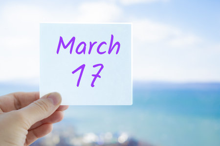 March 17th. Hand holding sticker with text March 17 on the blurred background of the sea and sky. Copy space for text. Spring month in calendar concept.の写真素材