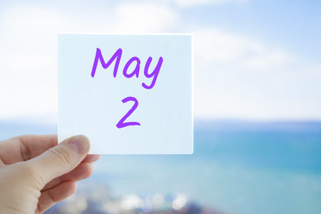 May 2nd. Hand holding sticker with text May 2 on the blurred background of the sea and sky. Copy space for text. Month in calendar concept.の写真素材
