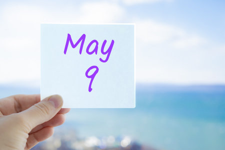 May 9th. Victory day. Hand holding sticker with text May 9 on the blurred background of the sea and sky. Copy space for text. Month in calendar concept.の写真素材