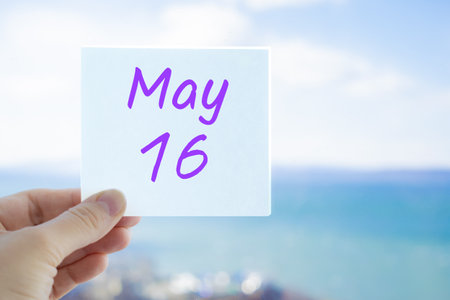 May 16th. Hand holding sticker with text May 16 on the blurred background of the sea and sky. Copy space for text. Month in calendar concept.の写真素材
