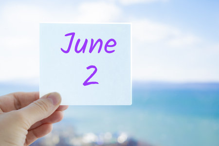 June 2nd. Hand holding sticker with text June 2 on the blurred background of the sea and sky. Copy space for text. Month in calendar concept.の写真素材