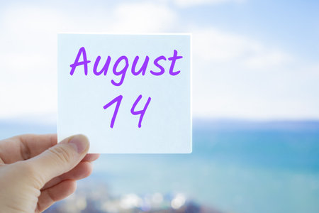 August 14th. Hand holding sticker with text August 14 on the blurred background of the sea and sky. Copy space for text. Month in calendar concept.の写真素材
