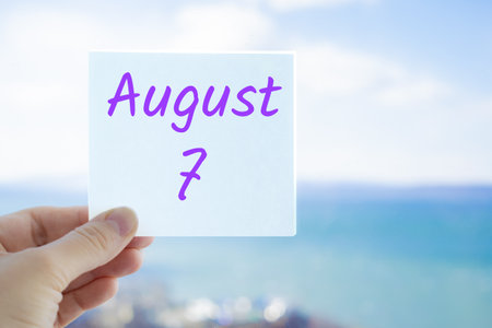 August 7th. Hand holding sticker with text August 7 on the blurred background of the sea and sky. Copy space for text. Month in calendar concept.の写真素材