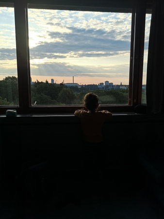 a little boy looks through a panoramic window at the city with green parks in the rays of the setting sun with rays breaking through the cloudsの写真素材