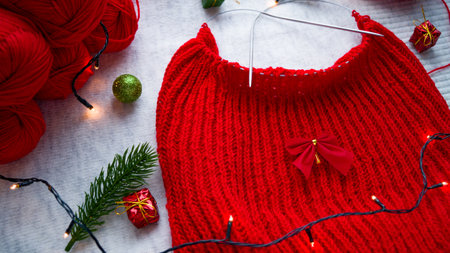 Knitting a red Christmas sweater on the background of New Years decor with a garland. Getting ready to celebrate Christmas and New Year. Knitting by hand. Woolen balls with red thread. Handmade.の写真素材