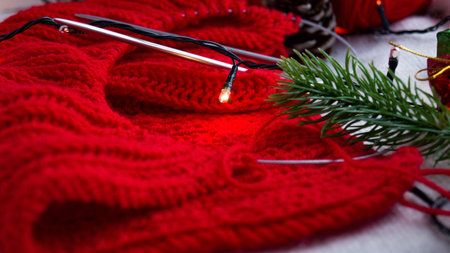 Knitted product of red color on hand knitting needles in the New Years decor, consisting of a garland and a spruce branch on a light background. Do-it-yourself knitting of a Christmas sweater.の写真素材