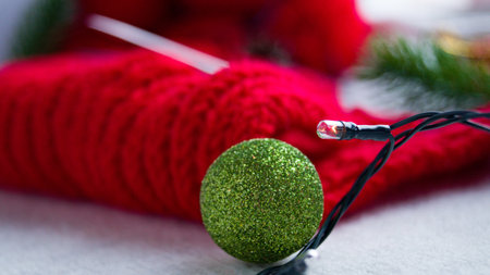 Green shiny ball for decorating the Christmas tree and New Year. On the background Knitted red product and a garland. Knitting a warm sweater for the winter. Hand knitting. Handmade.の写真素材