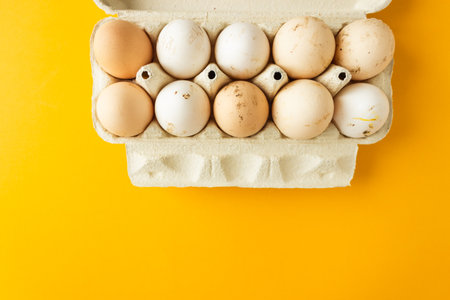 Top view of open egg box with ten brown and white eggs isolated on yellow background. Fresh organic...