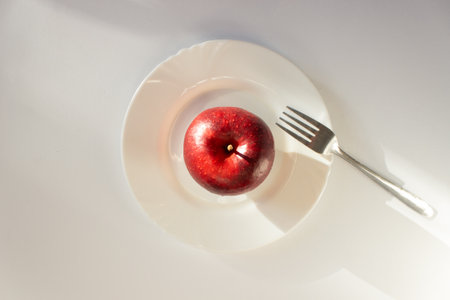 Whole ripe red apple on a white plate with fork. Isolated on white background. Ready to healthy eating. Raw food diet.の写真素材