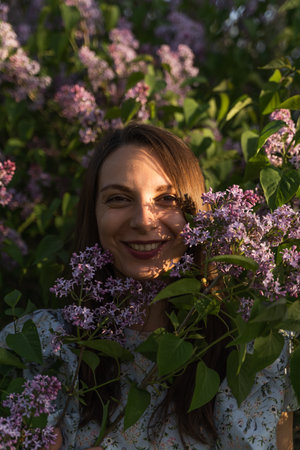 Portrait of attractive smiling caucasian woman surrounded by purple lilac flowers in the garden. Outdoor fashion photo.の写真素材