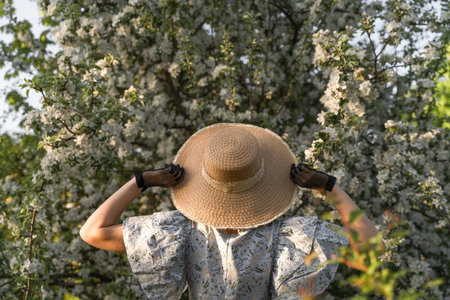 Young woman holds straw hat with her hands in black vintage lace gloves. Back view. Spring blossom...