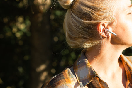 Young blonde caucasian woman listening music in wireless earphones on the city park. Cloe-up.
