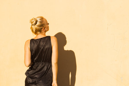 Beautiful tanned blonde in a black dress in bright sunlight isolated on a yellow wall background. Back view.の写真素材