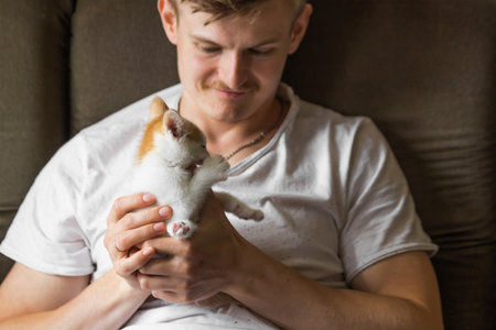 Young caucasian man in white t-shirt holding with hands a small white and red kitten at home. Man loves his pet.の写真素材